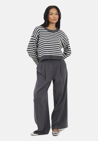 Vrouw in een grijs en wit gestreept trui, brede grijze broek en zilveren ballerina's, staand met haar handen achter haar rug.