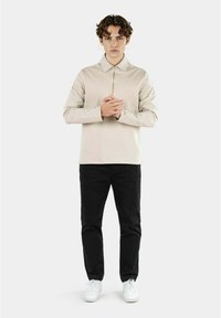 Camicia beige con zip e maniche lunghe, colletto, abbinata a pantaloni neri e sneakers bianche. Texture liscia, design minimalista.