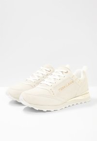 Baskets beige clair en suède, à bout arrondi, avec des lacets blancs, des accents dorés et une semelle en caoutchouc texturé. Marque Versace Jeans sur le côté.