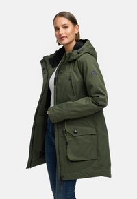 Frau, die eine grüne Kapuzenjacke mit mehreren Taschen trägt, seitlich steht und eine Hand in der Jackentasche hat, vor einem schlichten Hintergrund.