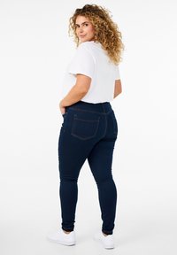 Jeans skinny de color azul oscuro con bolsillos y costuras en naranja, combinados con una camiseta blanca simple y zapatillas blancas. Pelo rizado visible.