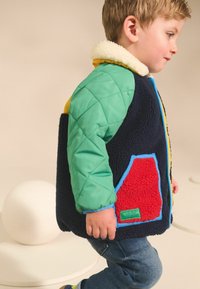 Veste colorée matelassée avec des manches vertes, un corps marine, une poche d'accent rouge et un col en fourrure légère. Détails cousus à la machine visibles.