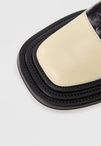 Sandale en cuir crème et noir avec un bout carré, un design superposé et une couture texturée le long des bords. Accentuée d'une finition brillante.