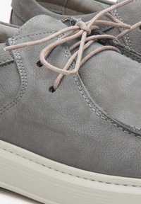 Chaussure en cuir gris au toucher lisse, avec un bout rond, une semelle blanche et des lacets gris clair noués en nœud. Des coutures sont visibles.