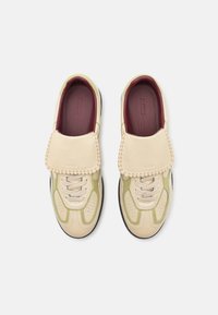 Baskets en cuir beige clair et vert avec fermeture à lacets, rabat en cuir tan rembourré et semelles texturées. Détail de couture notable sur le rabat.