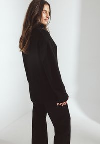 Juoda per didelė megzta suknelė su raukšlėmis ant rankovių ir apatinės dalies, turinti aukštą kaklą ir ilgas rankoves, derinama su juodomis plačiomis kelnėmis.