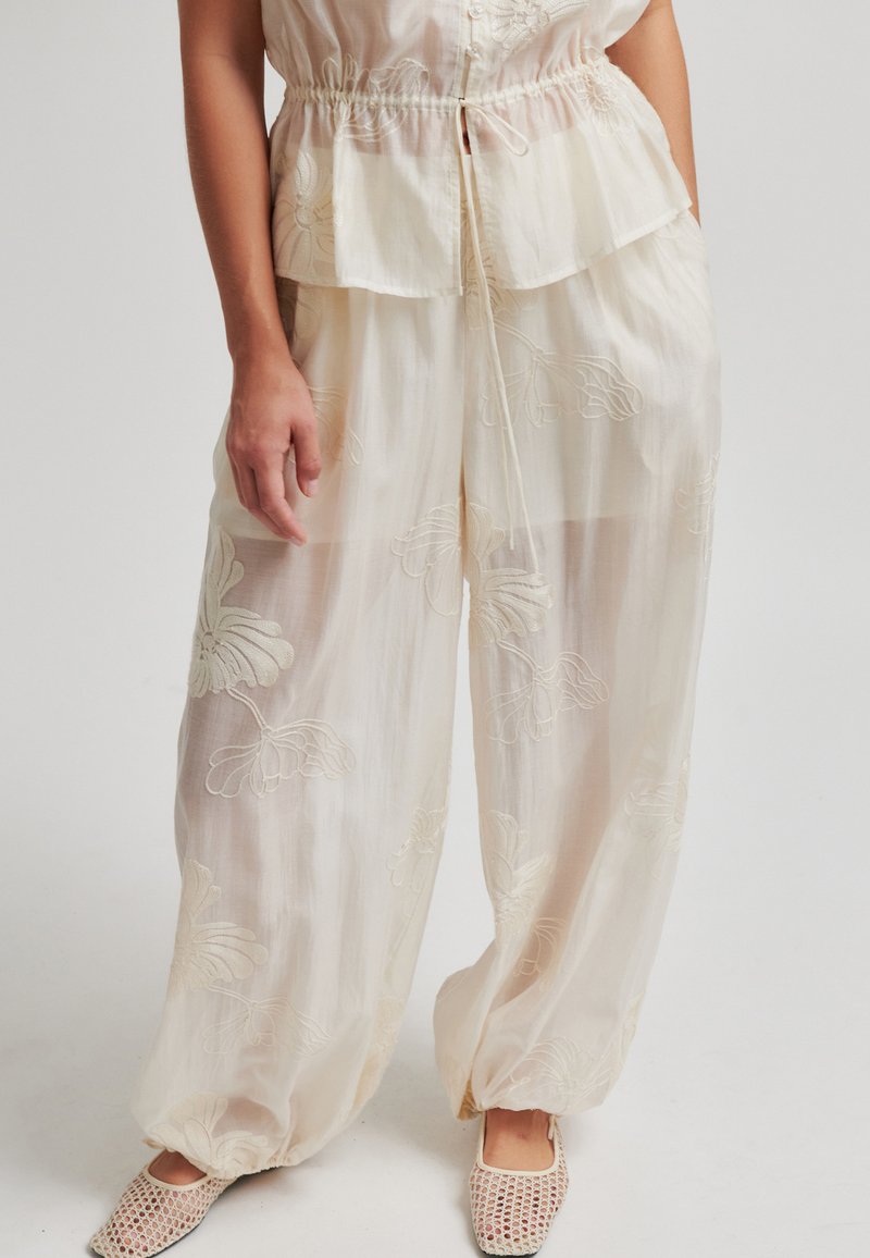 Femme portant un pantalon ample et transparent couleur crème avec une broderie florale, associé à un haut assorti et des chaussures plates tissées beiges.