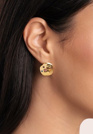 Gros plan d'une oreille avec une boucle d'oreille ronde en or ornée de petites pierres précieuses colorées en forme d'étoile.