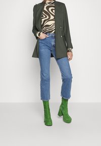 Grüne Wildleder-Stiefeletten mit dickem Absatz, blaue gerade Jeans, beige und braun gestreiftes Top mit Wellenmuster, grüne Doppelreiher-Blazer.