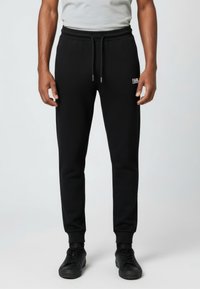 Pantalones jogger negros con cintura elástica, cordón ajustable, pierna cónica y pequeño logo blanco. Material liso sin patrones visibles.