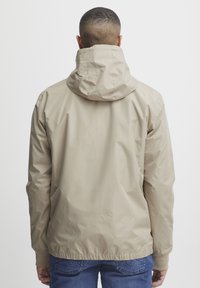 Beige nylonjakke med hette, elastiske mansjetter og nederkant. Glatt tekstur, minimalistisk design, ingen synlige logoer eller merker. Bakview vist.