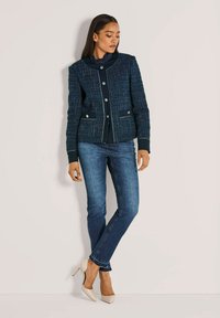 Frau steht mit überkreuzten Beinen, trägt eine strukturierte marineblaue Knopfjacke, dunkle Jeans und beige High-Heels vor einem einfarbigen Hintergrund.