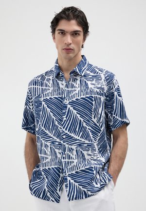 Chemise à manches courtes, à boutons, en blanc et bleu, avec un motif géométrique de vagues, col classique, et coupe décontractée. Matériau en coton.