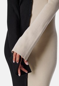 Deux mains avec des ongles rose clair, l'une dans une manche côtelée noire et l'autre dans une manche côtelée beige, se touchant doucement sur un fond uni.