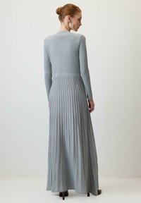 Kvinde med rødt hår i en knold, iført en lang, lys grå ribbet strikkjole og brune hælede sko, stående foran en ensfarvet hvid baggrund.