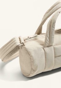 Jasnobeżowa prostokątna torba z wyściełanymi uchwytami i zapięciem na zamek. Wykonana z gładkiego, teksturowanego materiału, ozdobiona metalową plakietką z logo.