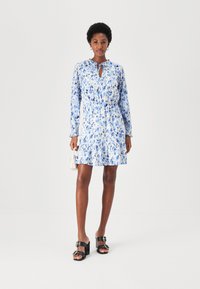 Blau und weißes Blumenkleid mit langen Ärmeln, elastischem Bund, Keyhole-Ausschnitt und Rüschensaum. Kombiniert mit schwarzen, hohen Sandalen.