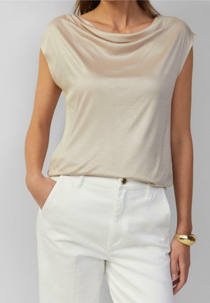 Žena v béžovém vršku bez rukávů s drapérií, bílých kalhotách s vysokým pasem a masivním zlatým náramkem, s jednou rukou v kapse.