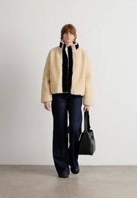 Beige scheerling jack met cropped mouwen over een zwart button-down shirt, gecombineerd met donkere flare jeans en een zwarte tote bag.