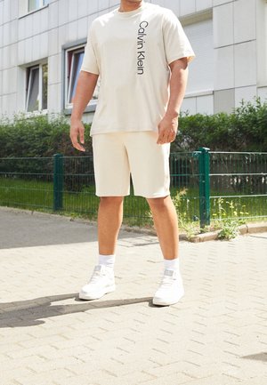 Homme portant un t-shirt crème Calvin Klein, un short assorti, des chaussettes blanches et des baskets blanches, debout sur un trottoir pavé en extérieur.