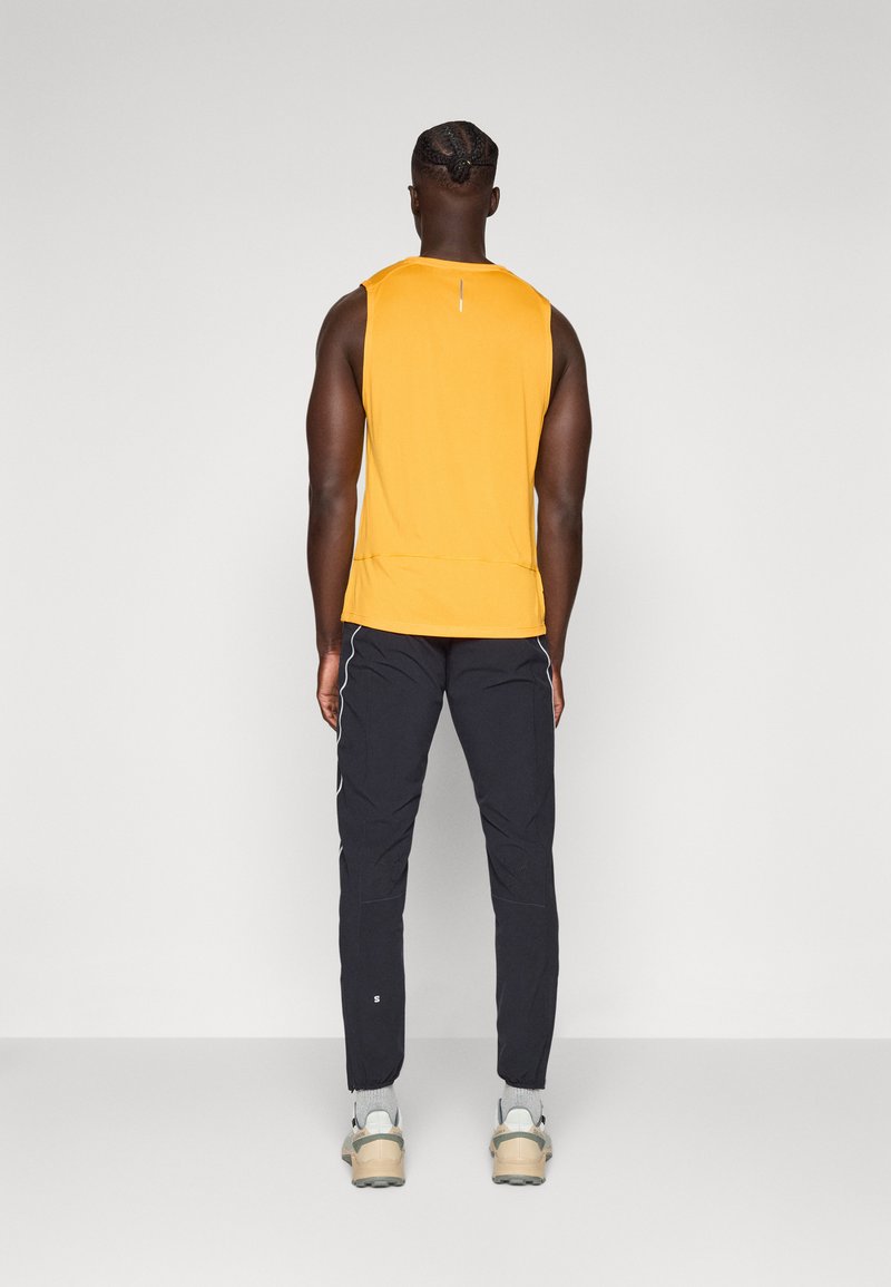 Homme se tenant de dos, portant un débardeur jaune, un pantalon de sport noir, des chaussettes grises et des baskets beiges sur un fond uni.