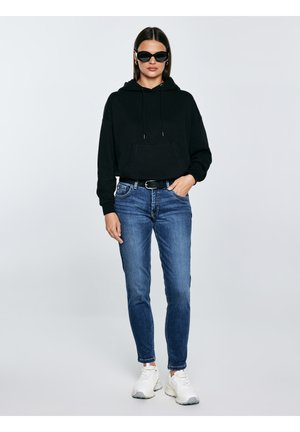 Nő áll, fekete kapucnis pulóvert, kék farmert fekete övvel, fehér tornacipőt és fekete napszemüveget visel, egyszínű világos háttér előtt.