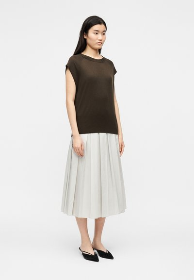 Femme aux longs cheveux foncés portant un haut marron sans manches, une jupe midi plissée blanche et des ballerines plates noires à brides arrière pointues, debout sur fond blanc.