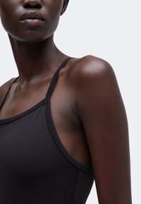Gros plan d'une personne à la peau foncée portant un haut noir sans manches, montrant son épaule, son cou et une partie du visage avec une expression neutre.