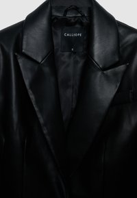 Veste en cuir noir avec revers aiguisés, une seule poche poitrine, et une étiquette indiquant "CALLIOPE", taille S. Texture lisse avec une coupe ajustée.