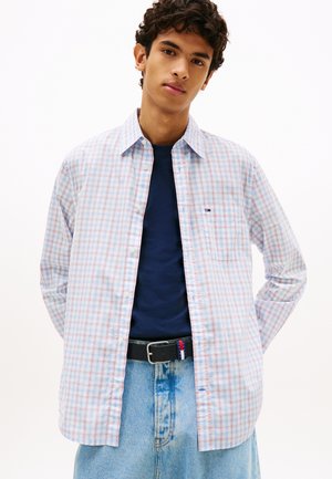 Młody mężczyzna ubrany w jasnoniebieską koszulę w czerwoną kratę na granatowej koszulce, jasnoniebieskie dżinsy oraz czarny pasek z klamrą w kolorach czerwonym, białym i niebieskim.