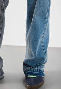 Nahaufnahme von Beinen, die ausgefranste blaue Jeans und marineblaue Sneakers mit grünen Schnürsenkeln auf einem schlichten hellen Hintergrund tragen.