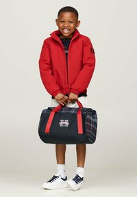 Giacca a vento rossa, maglietta nera, pantaloni corti bianchi e scarpe da ginnastica bianche. Tiene una borsa a tracolla a quadri neri con manici rossi e dettagli con logo.