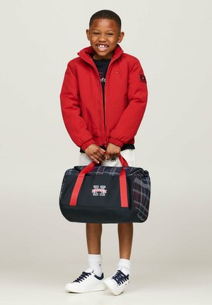 Giacca a vento rossa, maglietta nera, pantaloni corti bianchi e scarpe da ginnastica bianche. Tiene una borsa a tracolla a quadri neri con manici rossi e dettagli con logo.