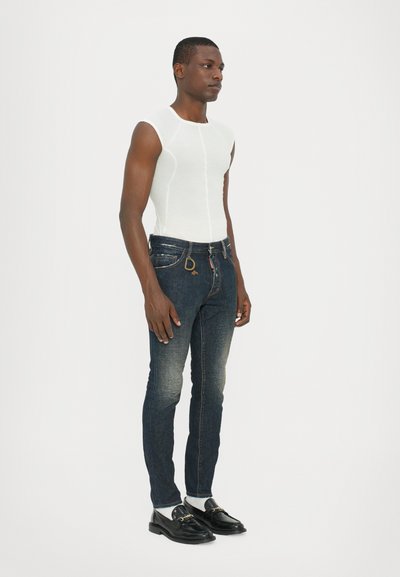 White sleeveless top, dark blue jeans with light fading, black shoes with gold accent, standing pose against a plain white background.