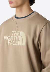 Beige sweatshirt med en tekstureret crew neck, der har et præget logo i en lysere nuance på brystet. Blødt stof med en afslappet pasform.