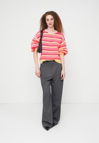 Πλεκτό πουλόβερ με πολύχρωμες ρίγες σε ροζ, πορτοκαλί και κίτρινο, συνδυασμένο με γκρι παντελόνι μεγάλης φαρδίας με ψηλή μέση και μαύρη τσάντα με πουά πουά σχέδιο.