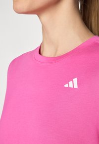 Vaaleanpunainen lyhythihainen paita, jossa on pyöreä pääntie sekä valkoinen logo, jossa on kolme raitaa vasemmassa yläkulmassa. Silkkinen, pehmeä kankaan rakenne.