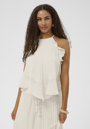 Femme aux longs cheveux bouclés portant une robe blanche sans manches à volants et des boucles d'oreilles créoles dorées sur un fond uni.