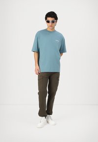 T-shirt en coton bleu ample à manches courtes, porté avec un pantalon cargo olive doté de poches latérales, et des baskets blanches.
