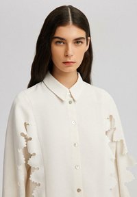 Blusa bianca con colletto e chiusura a bottone, caratterizzata da motivi floreali intagliati sulle maniche e un tessuto liscio e strutturato.