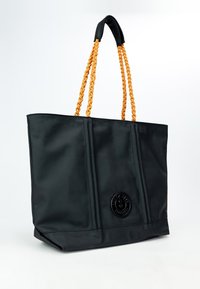 Czarna torba na ramię wykonana z gładkiego materiału, z plecionymi pomarańczowymi uchwytami, okrągłym logo na środku oraz usztywnionymi bokami z przeszyciami.