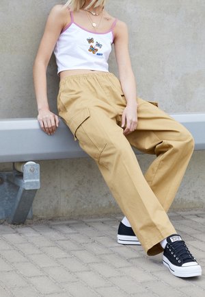 Młoda osoba siedzi na szarej ławce, ubrana w beżowe spodnie typu cargo, czarne sportowe buty i biały krótki top z motywem motyli.