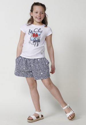 Camiseta blanca con diseño gráfico azul y rojo, combinada con pantalones cortos navy con patrón. Los zapatos son blancos, con una correa y suela de cuña.