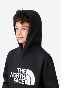 Črna jopa iz mehke tkanine z velikim belim logotipom "THE NORTH FACE" na sprednji strani, z zategovalno kapuco in rebrastimi manšetami.