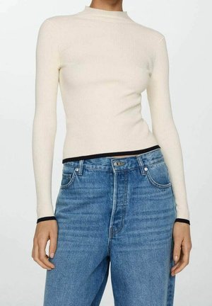 Strakzittende, lange mouw beige geribbelde trui met zwarte rand bij de manchetten en zoom, gecombineerd met een high-waist blauwe spijkerbroek.