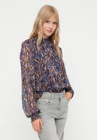 Mujer con cabello largo y rubio que lleva una blusa de cuello alto y mangas largas con patrones abstractos en azul y marrón, combinada con unos jeans gris claro.