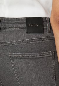 Vaqueros de mezclilla gris oscuro con un parche de cuero etiquetado "BOSS" en la cintura, bolsillos traseros visibles y detalles de costura.