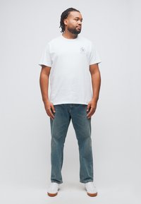 Άνδρας με πλεγμένα μαλλιά, φοράει λευκό T-shirt με σχέδιο, μπλε τζιν και λευκά αθλητικά παπούτσια, στέκεται και κοιτάζει προς τα δεξιά του.