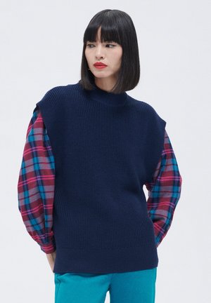 Maglione blu lavorato a maglia senza maniche con collo alto, indossato su maniche lunghe a quadri multicolori. Presenta una vestibilità comoda e un materiale testurizzato.