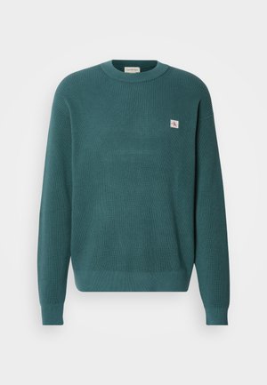 Tyrkisk teksturert genser med ribbestrikket mønster, rund halslinning og lange ermer. Har en liten brodert logo på brystet.